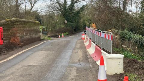 Cones, bollards and fencing along the right hand side of the bridge. Some brickwork damage can be seen. A road going through centre of image. On the left is the undamaged side of the bridge.