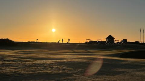 Golfers silhouetted on the course against an orange sunrise glow.
