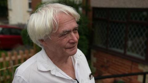 A man with white hair wearing a white shirt, standing in his garden.
