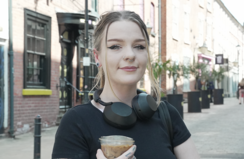 Young woman looking to camera in a street in Wolverhampton. She's holding a coffee and has headpones around her neck. She's wearing a nose peircing and has brown hair, black top. 