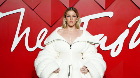 Ellie Goulding wearing an off the shoulder white winter coat. She has dark blonde hair that goes down her back and silver earrings. She is stood in front of a red background with white wording across it.