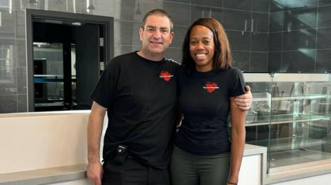 A man and a woman both wearing black T-shirts and black trousers look smiling at the camera with the backdrop of an empty cafe behind them. The man has has left arm around the woman's shoulder.