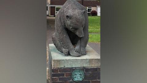 A sculpture of a bear in a square in the centre of houses. The sculpture sits on a brick plinth and there is a metal plaque attached.