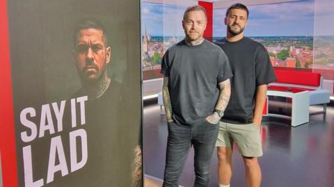 On the left, Danny Tanti, who has short black hair combed forward, and a black beard and moustache. Jonny Human, on the right, has short dark hair combed up on top and cropped short on the sides. He has a light brown full beard and moustache. They are both wearing a Say It Lad branded T- shirt, and are standing inside the BBC Look East studio, with their logo and an image of Jonny on the tower screen.