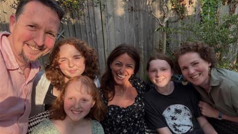 Six people squeezing in for a selfie. Davina McCall is in the middle, she is surrounded by three young girls and their mum and dad. They are sitting in a garden.