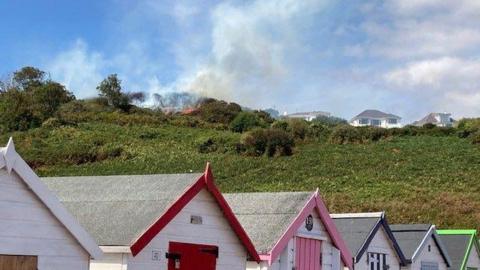 There is a fire at the top of the hill. Along the bottom are beach huts which are different colours. There are also houses at the top of the hill on the right.