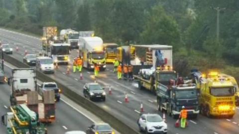 A CCTV image of the section of the M25. A single-file line of traffic is passing through the single open lane. Next to it are several large trucks and people in high-vis.