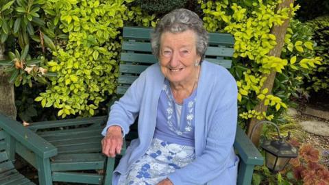 Ann Lush sat in her garden on a green wooden chair. Next to her is an empty matching chair. She is an elderly woman wearing a blue and white floral dress, with a matching blue cardigan. She is smiling at the camera