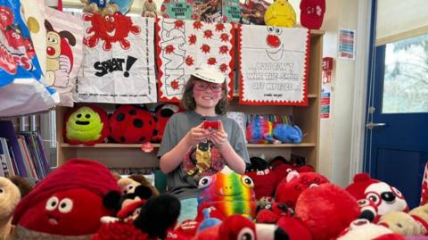 Ziggy wearing big red glasses and a white cap shows off his massive collection of Comic Relief items from across the decades. The picture shows an array of soft toys, tea towels and tote bags.