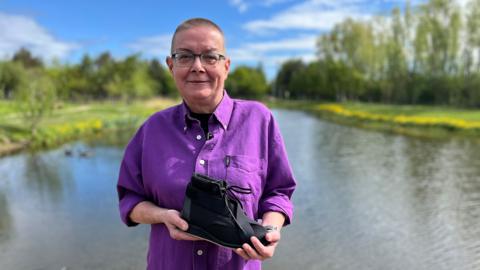 Jeanette Russell smiles and she stands in front of a body of water. It's a sunny day and there is lush green grass on either side with a row of trees and hedges on either side. She has cropped hair and is wearing glasses and a bright purple shirt. She is holding the black lace-up shoe.