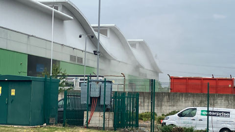 A general view from the road of a large white, green and brown industrial building behind a green fence. Vehicles are parked inside the fence and white smoke can be seen coming from the premises.