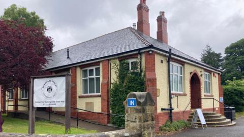 The exterior of a small, single-storey community building situated in a quiet, green area. The building is constructed with cream-coloured textured walls and red brick detailing around the corners, windows, and doorways. The roof is covered with dark grey slate tiles and features multiple brick chimneys rising above it.
On the left side of the image, a large sign is mounted on a wooden frame. The sign clearly reads: “Newby and Scalby Community Hall” and underneath it says: “Contact for Bookings and Event Hire 01723 500699".