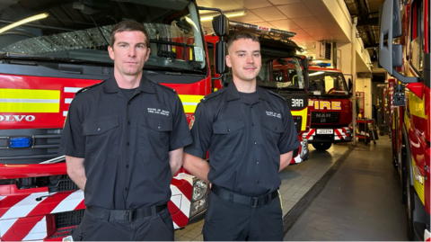 Toby Edwards and Logan Le Flem stand side by side at a fire station. The pair are dressed in uniform; black shirts and black trousers. They both have white skin and brown hair. They are stood in front of a number of fire engines with their arms behind their backs. They are looking at the camera. 