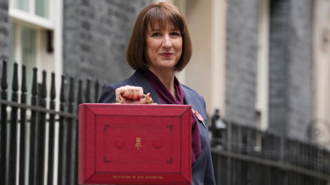 Rachel Reeves, a woman with dark brown hair, dressed in a grey suit with purple scarf, holds up a red box with gold coloured handle and gold coloured lettering in street with black railings behind her.