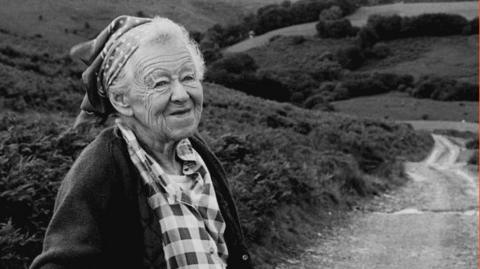 A black and white photograph of an elderly lady with white hair wearing a head scarf, a checked blouse and cardigan, with a road through Exmoor behind her