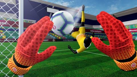 A screenshot of the virtual reality game, the image shows a pair or red goalkeeper's gloved hands from the perspective of the player. A second player in a yellow kit is kicking a white ball towards a goal. They are in a virtual. empty stadium.