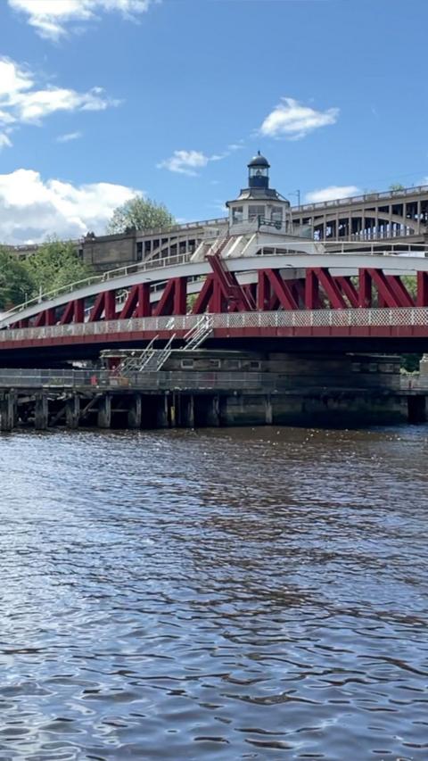 A red-and-white metal swing bridge spans a river, viewed from the water’s edge. The bridge features a small central control tower with a domed roof and glass windows. A staircase leads up to the bridge deck, and a raised roadway runs behind it. The foreground shows rippling water, while the background includes green trees and a blue sky with scattered white clouds.