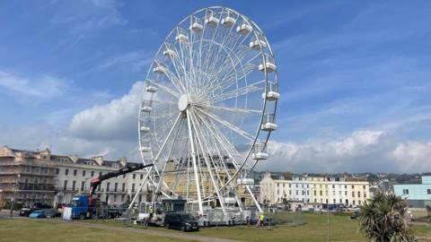 The wheel is white and looks large. It is set in a grassy square lined with houses. A lorry has a extendable metal arm leaning towards the wheel and the base is surrounded by metal fencing.