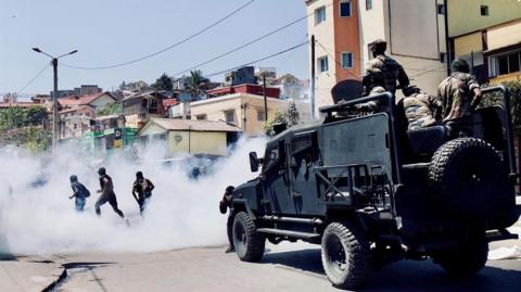 Riot Police in a heavily armoured bear-cat kind of vehicle drive behind three protesters who are running away through tear gas smoke.