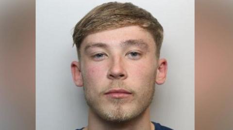 A young white man with blue eyes, mousy- blond hair and stubble. He is wearing a navy top with a round neck