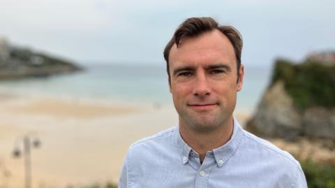 A man stands in front of a beach in Newquay. He is wearing a blue shirt. 