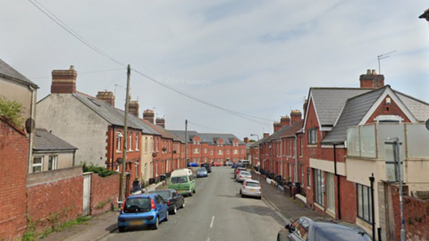 Google map street view of Bruce Street, Cathays, Cardiff. Red brick terrace houses line either side of the street. Cars are parked either sides of the pavements. The sky is grey. 