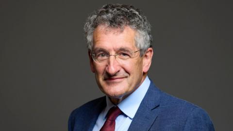 Peter Pinsley is in the middle of the picture staring at the camera smiling. He has dark grey hair, round glasses, and is wearing a blue shirt and suit jacket with a red tie. He is standing in front of a grey background. 