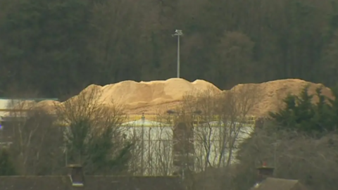 Photograph of Kronospan Limited in Wrexham. Mountains of sand can be seen in the distance, behind some leafless trees. A lampost can be seen in the far background, as well as large dark pine trees.