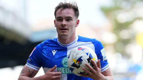 Kofi Balmer holds a football in his right hand while on the pitch during a match for Bristol Rovers this season