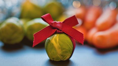 A Brussel sprout wrapped in a ribbon with a bow on top on a dark surface. Other sprouts and carrots are piled behind but are out of focus.