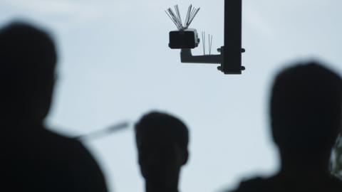 Silouettes of three people and a CCTV camera on a pole - the cameras has spikes on the top of it