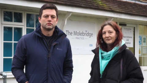 Chris Coghlan MP, left, wearing a navy blue coat, and Caroline Joseph, right, wearing a light blue hoodie and a black coat. They are stood in front of The Molebridge Practice in Fetcham, a white building with a sign reading "The Molebridge Practice" on the front.