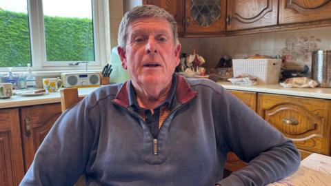 Gerry White sits at his kitchen table, he has short grey hair and is wearing a blue quarter-ziip top with maroon detailing on the inside of the collar. Behind him are brown wood kitchen cabinets and items including a radio, block of knives and bread basket on top of the kitchen counters.