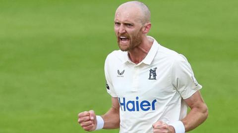 Olly Hannon-Dalby pumps both fists by his side in celebration after taking a wicket for Warwickshire. He has a shaved head and closely clipped bears and is shouting.