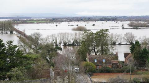 A number of flooded fields are seen from above. Two houses are visible in the foreground, surrounded by water.