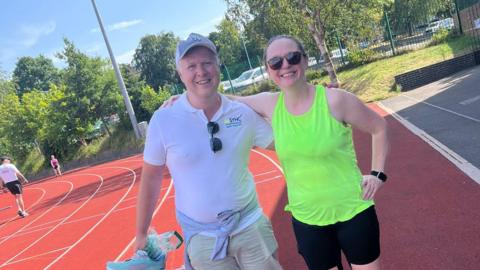 Sarah Dodd standing on the track of Wentworth Leisure Centre after her fundraising challenge. She is wearing her running clothes, including a bright green vest. She has her arms around a man, wearing a white shirt and cap.