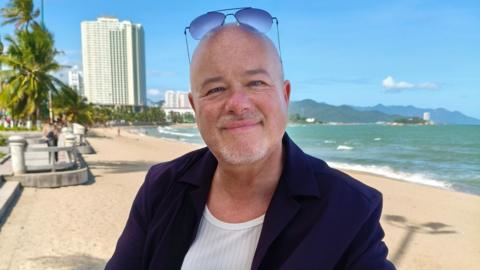 Man with bald head and sunglasses, smiling at the camera while standing on a beach with the sea lapping up behind him