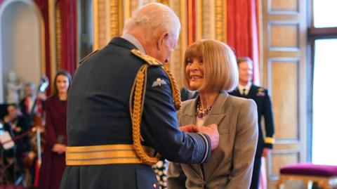 King Charles pins the order of the Companion of Honour onto Anna Wintour's grey lapel. She is smiling, and not wearing her sunglasses 