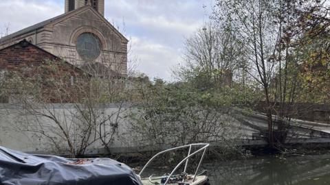 A view of the derelict site with tattered banners alongside the canal in Jericho. The church can be seen in the background. There is also a boat in the water.