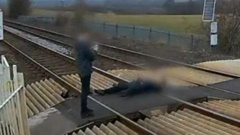 A blurred CCTV image shows a boy or man standing up taking a photo of another boy or man who is lying down on railway tracks at a level crossing. Their faces are blurred on purpose.
