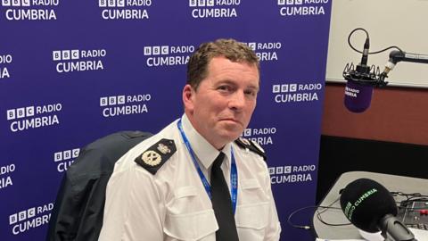 Chief Constable Rob Carden sitting behind a microphone in the BBC Radio Cumbria studio. He is wearing his police uniform.