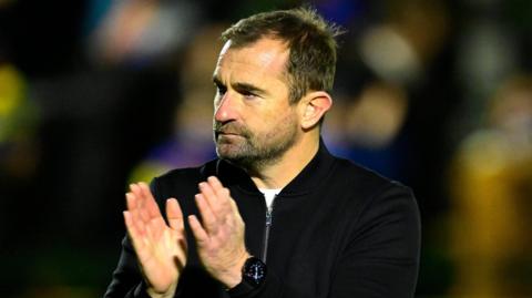 Paul Wotton, wearing a white t-shirt beneath a dark sweater, applauds the football fans at the end of a match