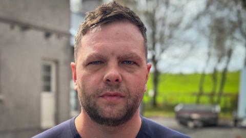 A man in a T-shirt frowning at the camera, with a fluffy microphone close to his face. A house, some trees and a green field are visible behind him.