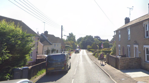 A general view of the road through Dennington. Houses line either side of the road as vehicles can be seen travelling down. On the right is a woman walking her dog along a pavement. 