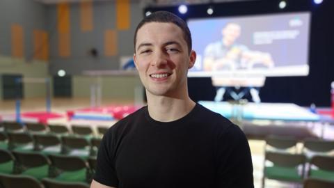 Rhys McClenaghan. He has short dark hair, wearing a black t-shirt and smiling at the camera. Behind him are rows of chairs and a large screen.