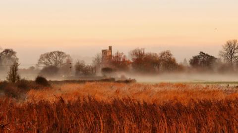 A church sits on the horizon shrouded in mist. There are also trees either side of the church. In the foreground, there is a field of wheat almost orange in the early morning light.