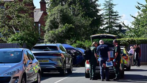 There is a ambulance car with paramedics. One is holding a defibulator.
To the left is a silver car. Behind that is a blue car crashed into the hedge. The car is tilted to its side.