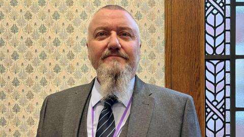 Councillor James Bean. He is looking at the camera. He has short hair, a long grey beard. He is wearing a light blue shirt, a dark blue tie and a grey jacket. 