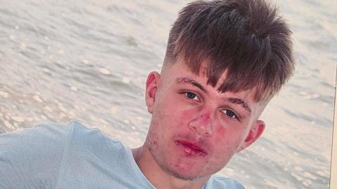 An image of a boy with medium length brown hair looking into the camera. He is wearing a light blue t-shirt and is in front of the water