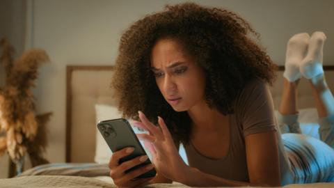 Woman with brown hair looks concerned while laying on a bed and looking at her smart phone